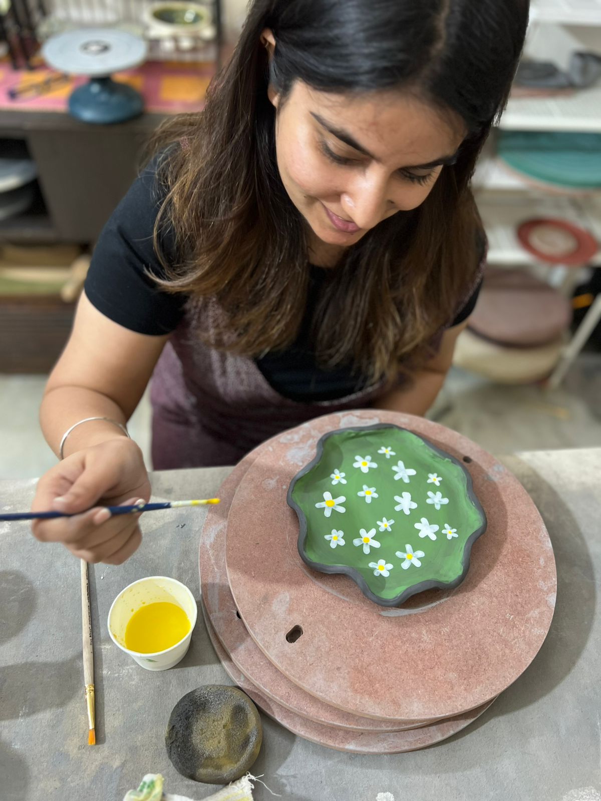 Woman smiling while painting white flowers on a green pottery plate using a fine brush.